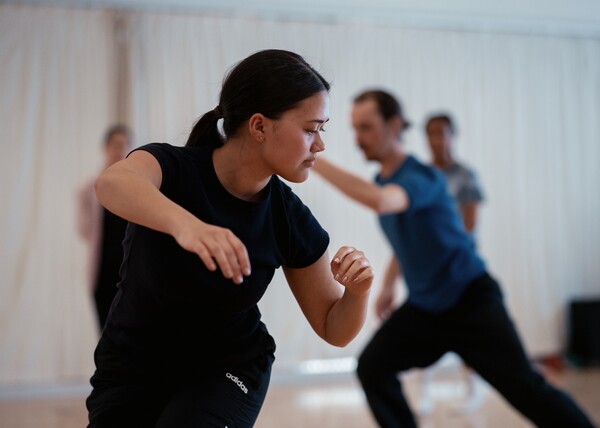 A person in the forefront of the image is wearing a black t-shirt and trousers. They are standing, knees bent, and their body is twisted as they look to the left towards the floor. Their right arm reaches forward with a bent elbow, palm facing the floor. The left arm is bent by the side of their body.  In the background here are three other dancers in the studio