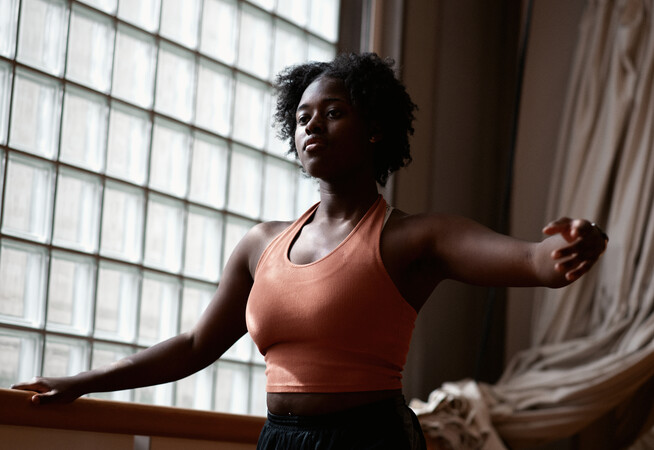 A person wearing a light orange halterneck top and black bottoms. Their right hand is placed on a ballet bar, and their left arm is outstretched to the side of their body angled forward, elbow slightly bent and palm facing forward.