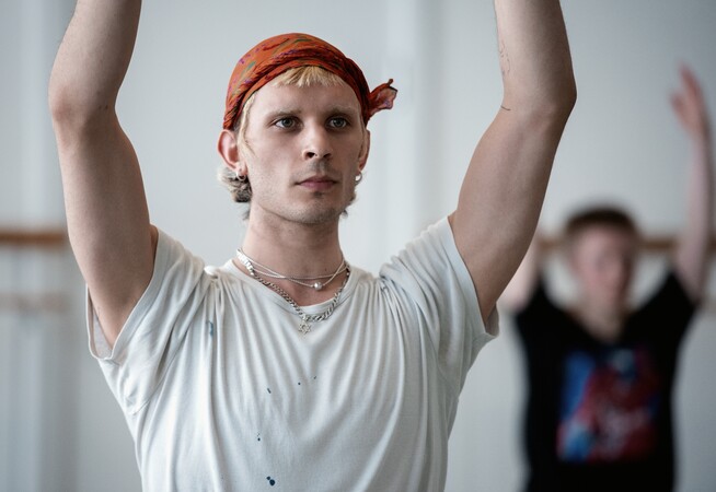 In a dance studio, a dancer in a white t-shirt with an orange scarf around their head stands with both arms reaching upwards, elbows slightly bent. A dancer in the background stands in the same position, arms up and elbows bent with palms facing towards each other. They wear a black t-shirt.