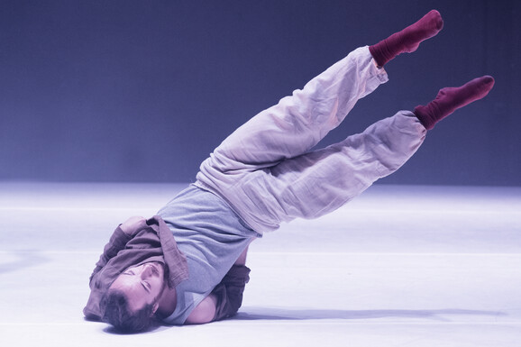 A dancer wears a grey vest, brown shirt, beige cargo trousers and deep red socks. They are on stage lit by a blueish wash. Their upper body is on the floor, their arms behind their torso with elbows bent, arms support their back. Their lower body is lifted off the floor and points diagonally upwards so that their whole body makes a diagonal line from floor up to the right of the image.