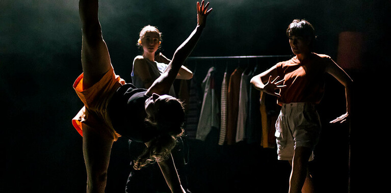 A dancer stands with one leg on a dark stage, her other in a straight line above her in the splits. Her torso is parallel to the floor, her arms outstretched. Behind her are two other dancers, back lit by a high contrast light, shining through haze.