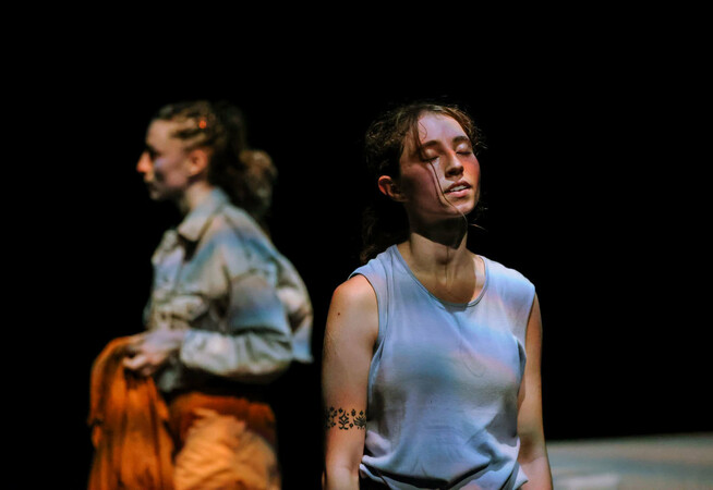 A dancer in a blue knitted vest rests for a moment, her eyes closed, a slight smile on her face. Perspiration on her face suggests she's just been dancing. Behind her, and out of focus, another dancer carries a jacket, walking with purpose across the stage.