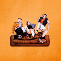 Three dancers leap up onto a brown leather sofa, smiling big warm smiles at each other, clearly they are close friends. Their outfits are all 90s inspired in shades of orange and red. The background of the image is an orange, textured like paper.