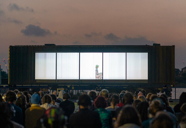 Set against the sky at dusk, a captive audience all wearing headphones are seated in front of a large, dark shipping container, mounted on the bed of a 40ft truck. The long side of the container has been removed revealing a single silver being, crouched on the floor.