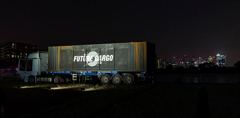 A shipping container, mounted on the back of a 40ft truck, has the words Future Cargo written in bold, white writing on the long side. In the distance, behind the container are the bright lights of a city skyline at night.