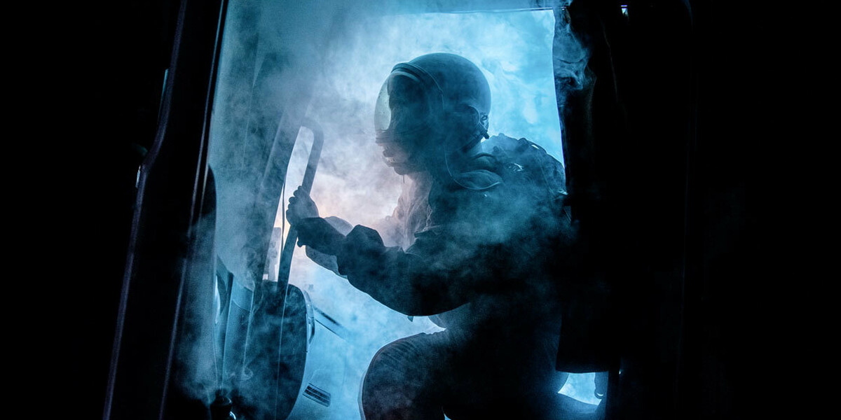 The cabin door of a truck opens, spewing eerie blue smoke. Silhouetted by a bright blue light that cuts through the darkness of night, a figure wearing an astronaut uniform begins to emerge.