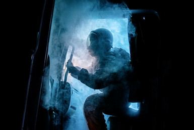 The cabin door of a truck opens, spewing eerie blue smoke. Silhouetted by a bright blue light that cuts through the darkness of night, a figure wearing an astronaut uniform begins to emerge.