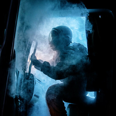 The cabin door of a truck opens, spewing eerie blue smoke. Silhouetted by a bright blue light that cuts through the darkness of night, a figure wearing an astronaut uniform begins to emerge.