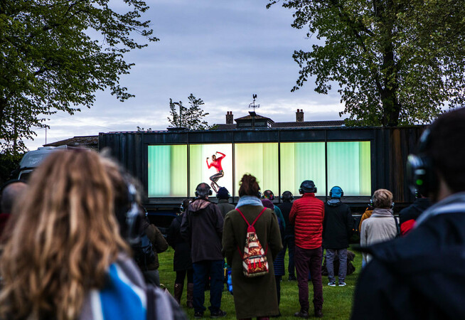 An audience of people are stood facing a shipping container, all wearing headphones. The long side of the shipping container is open, revealing a shiny silver being dancing in a neon pink jumper. The inside of the container is lit up in bright green.