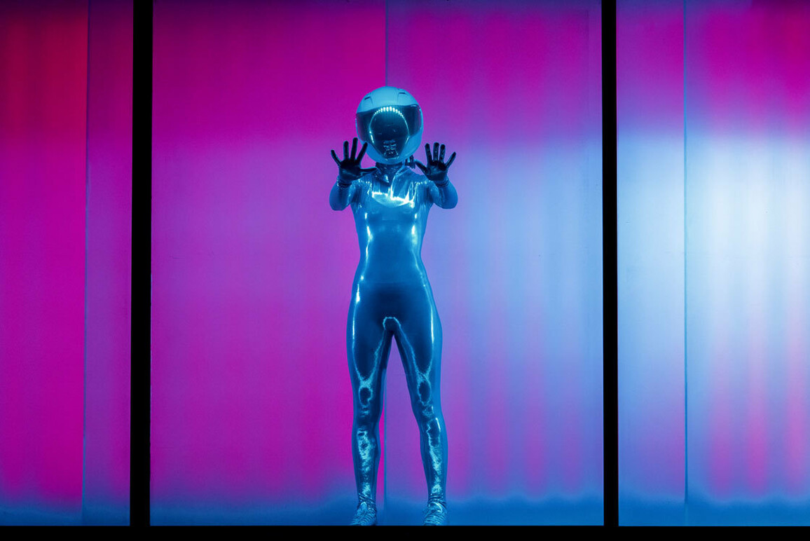 A shiny silver being extends their hands out towards us, placing their hands on an invisible pane of glass that separates the world inside the shipping container, lit up in contrasting shades of red and blue, and our world. They are wearing a protective helmet with a mirrored visor. Their head is cocked inquisitively to one side as they gaze towards us.