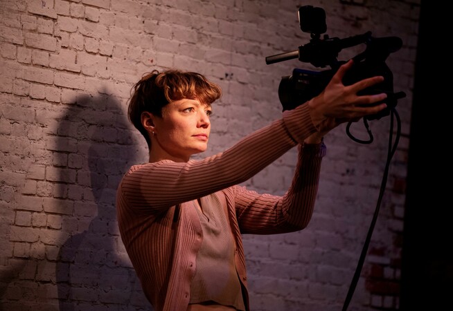 A female performer is standing if front of a white bricked wall holding a camera which she has turned in her direction facing her and lighting her face.