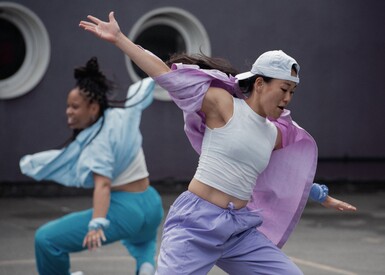 Two performers are seen dancing outside. One is wearing pastel purple shirt and trousers with a white vert and a white cap, the other, behind her is wearing matching pastel blue shirt and trousers and a vest