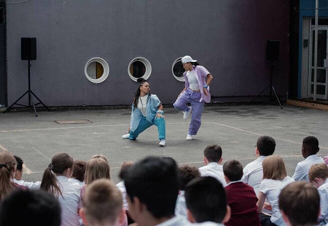 Two dancers dance together in a playground, one wears blue trousers and an open blue shirt over a white t-shirt, and the other has purple trousers and a similar purple shirt. In the foreground, there is a large crowd of young school kids all watching intently.