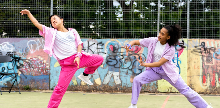 Lisa, a Southeast Asian, female dancer of small build, with black shoulder-length hair and black eyes, dances on the left with her left arm raised and her eyes gazing towards it. She is wearing a white shirt, dark pink trousers and a light pink button-up shirt, with a pink bandana around her wrist. Kassi, who is taller and has brown skin and afro-curly hair, lunges towards Lisa with arms outstretched. Kassi is wearing light purple trousers, a light purple shirt with a white vest underneath. She is wearing white trainers.