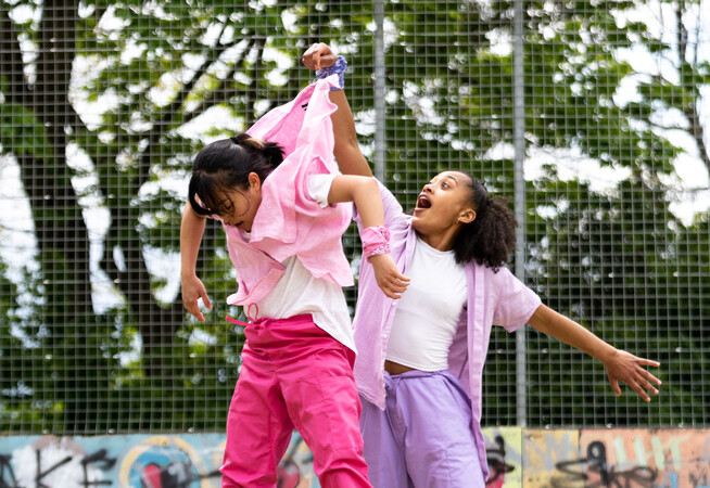Lisa, a Southeast Asian, female dancer of small build, with black shoulder-length hair and black eyes, is being thrown in the air by the scruff of her neck by Kassi who is much taller! Lisa is wearing a white shirt, dark pink trousers and a light pink button-up shirt, with a pink bandana around her wrist. Kassi, who has brown skin and afro-curly, is wearing light purple trousers, a light purple shirt with a white vest underneath. She is wearing white trainers.
