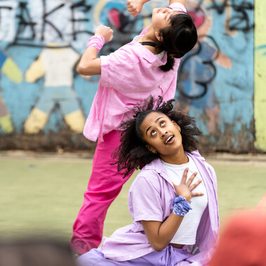 Lisa, a Southeast Asian, female dancer of small build, with black shoulder-length hair and black eyes, wearing a white shirt, dark pink trousers and a light pink button-up shirt, with a pink bandana around her wrist, dances with her back to the audience, her bending backwards with her fists at her chest and her elbows held high. Kassi, who has brown skin and afro-curly hair, wearing light purple trousers, a light purple shirt with a white vest underneath, is seated behind Lisa. Her hand is held to her chest, fingers splayed, her eyes looking up to the sky. The audience are visible in the foreground.