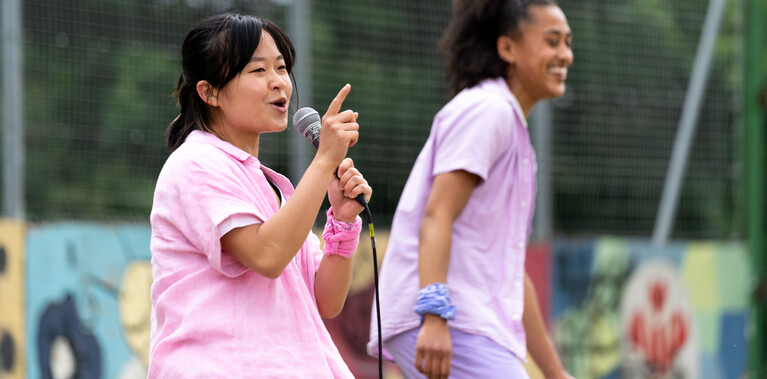 Lisa a Southeast Asian, female dancer of small build, with black shoulder-length hair and black eyes, wearing a white shirt, dark pink trousers and a light pink button-up shirt, with a pink bandana around her wrist, talks into a microphone and gestures with her figure. Kassi, who has brown skin and afro-curly hair, wearing light purple trousers, a light purple shirt with a white vest underneath, stands in background