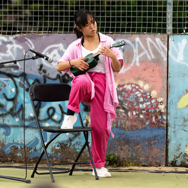 Lisa, a Southeast Asian, female dancer of small build, with black shoulder-length hair and black eyes, wearing a white shirt, dark pink trousers and a light pink button-up shirt, with a pink bandana around her wrist, plays a small green ukulele close to a microphone, one knee raised on a chair.