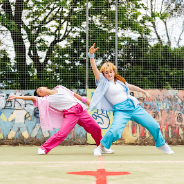 Lisa, a Southeast Asian, female dancer of small build, with black shoulder-length hair and black eyes, dances with her legs apart and her left arm and torso parallel to the ground. She is wearing a white shirt, dark pink trousers and a light pink button-up shirt, with a pink bandana around her wrist. Rose copies the shape of Lisa’s dance but has her hand pointed upwards. She has pale freckled skin, blue eyes, ginger curly hair, is small in height and wearing bright blue trousers with a pale blue shirt.