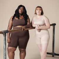 Two plus sized dancers wearing varying skin toned, body hugging shorts and bras, stand at a ballet bar. They stare out towards us, asking us to question what we see.