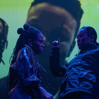 Yuki (who is East Asian, Japanese) and Sarah (who is mixed race, white British and west African, with long dark brown twists) groove intensely together, wearing matching black shiny outfits. In mid-motion, Yuki sways backwards to the left with one arm raised in the middle of the image, while Sarah leans towards them, gazing intently. Behind them, a music video is being projected with three singers looking down on them.