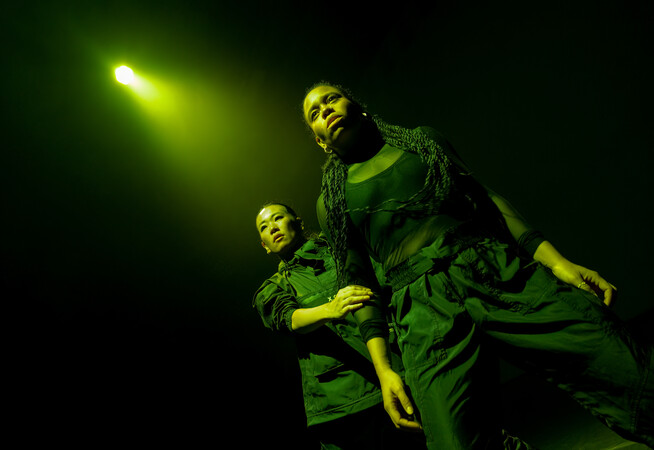 Sarah (who is mixed race, white British and west African, with long dark brown twists) stands with her legs apart and her gaze upwards. Yuki (who is East Asian, Japanese) grasps her arm and stands behind her, following her gaze. They are lit by a single green light that reflects off their shiny black outfits.