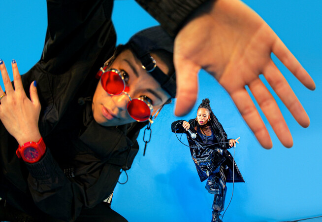 A dancer in all black trackies, a backwards black cap and red sunglasses and watch leads into the camera, their hand is up to the camera and is warped by a fish eye lense. In the background, another dancer in all black dances with a microphone in their hand. They slot into the space between the thumb and forefinger of the dancer in the foreground. The background is a bright electric blue.