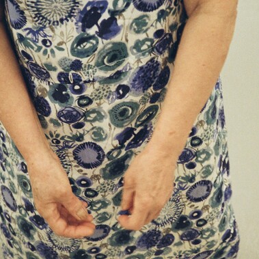 A close-up of an elderly woman’s arms and hands wearing a blue  floral patterned dress, with a focus on the intricate flower designs and the position of the hands in subtle motion.