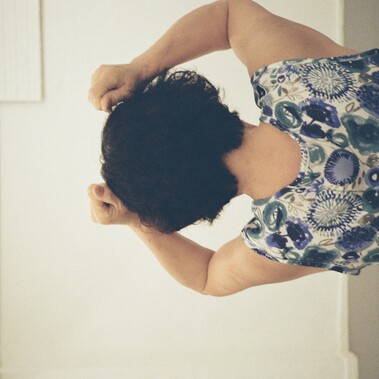 An elderly woman with short, dark and curly hair is seen from behind,  with both hands raised near their head while wearing a floral-patterned clothing