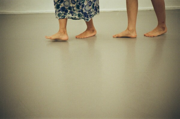 Two pairs of bare feet walking on a smooth floor, one set of feet  appears to belong to an older individual wearing a long flower patterned  garment, while the other set belongs to a younger person.