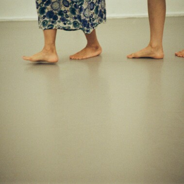 Two pairs of bare feet walking on a smooth floor, one set of feet  appears to belong to an older individual wearing a long flower patterned  garment, while the other set belongs to a younger person.