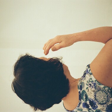 A elderly woman with short, dark curly hair wearing a floral patterned  clothing is seen from behind, raising her hand to her face in a thoughtful  gesture, against a plain background.