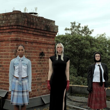 5 females stand in a line, looking strongly at the camera with a dead-pan expression and pose. All wear a mixture of textured and coloured clothing.