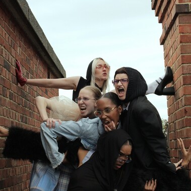 5 females are intertwined with animalistic facial expressions. All wear a mixture of textured and coloured clothing and stand between two brick structures.