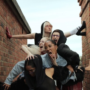 5 females are intertwined with animalistic facial expressions. All wear a mixture of textured and coloured clothing and stand between two brick structures.