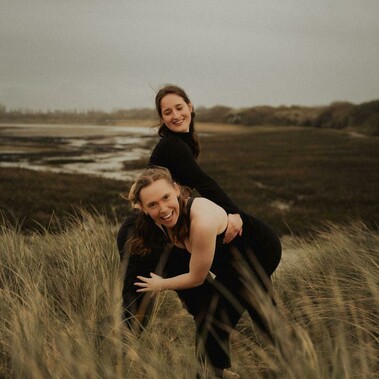 Two dancers with long hair, one leaning forward in the foreground, hold on to each other while smiling outside.