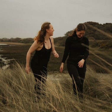 Two women, one with light brown hair and the other with dark brown hair, stand side by side laughing.
