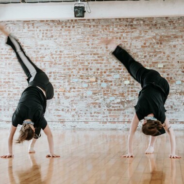Two dancers side by side stand inverted with one leg toward the ceiling slightly out to the side.