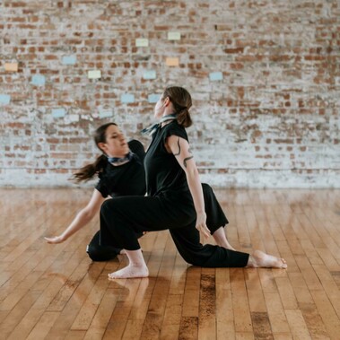One dancer in front kneels on the floor and another dancer behind sits on their knee leaning to the side.
