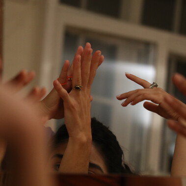 A square mirror that’s reflection holds the top of one woman’s head in the background, in-front of  a blurred white frame window. The foreground of the reflection holds feminine hands touching in  various ways. Two of the hands wear rings.