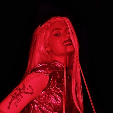 A young white woman with long blond hair wearing a silver bodice, wrapped in rope,  smiles menacingly at the camera while biting a piece of rope. The image has a red hue with a black background.