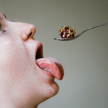 A glacé cherry covered with gold star pins is held out on a silver teaspoon. Below, a white person greedily sticks out their tongue, ready to devour.