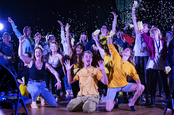 aKa Dance performers and the audience are all together in a large group, cheering and throwing confetti in the air. The performers are kneeling on the floor, whilst the audience are stood behind them.