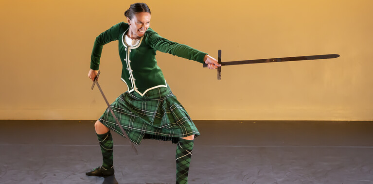 Charlotte Mclean, the performer, is photographed in green tartan kilt, knee-high socks, and green top. She is wielding two wooden swords and is in an offensive stance, with her legs firmly planted and open. She points one sword forward with her left arm as if stabbing are signalling others forward.