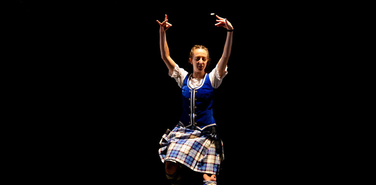 The performer, Charlotte Mclean, is photographed in a blue and white tartan kilt, knee-high socks, and blue vest. She is mid-dance, with her hands raised in the air and one leg firmly on the ground, whilst the other is slightly lifted.