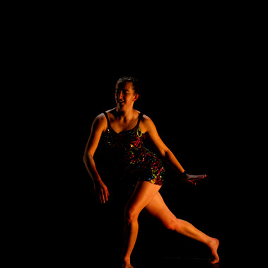 Charlotte Mclean, the performer, is photographed sidestepping along the stage. The backdrop and stage floor are pitch black.