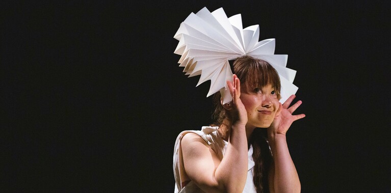 A performer holds a fanned paper circle around their head and smiles.