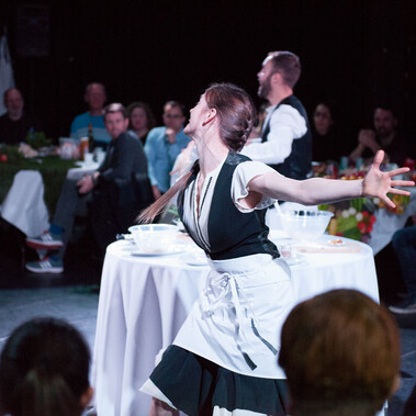 A performer, dressed as a waitress, is captured with their arms spread apart, gliding forward. Begin them, an audience seated at dinner tables looks on.