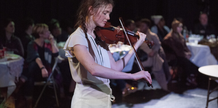 A female performer is captured playing the violin, their head bowed and eyes closed, as if lost in a moment of calm and focus. Begin them, and audience seated at dinner tables watches on.