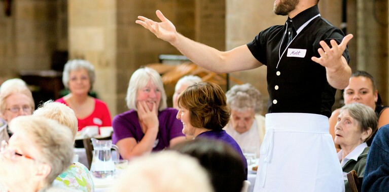 A performer, dressed as a waiter, stands with arms spread out, in the midst of either singing or talking with the audience. He is surrounded by an audience seated at tables, who look on with amusement.